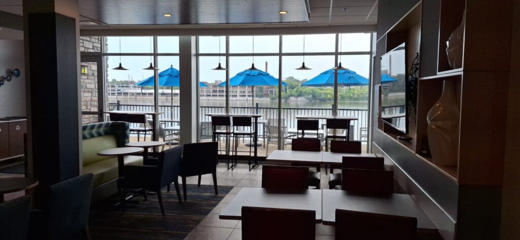 The image shows the interior of a restaurant or cafe with tables and chairs arranged near large windows. Outside, there is a patio area with more tables and chairs, shaded by blue umbrellas. The windows offer a view of a body of water and a distant shoreline with trees and buildings. The interior has a modern design with a mix of seating options and decorative elements.