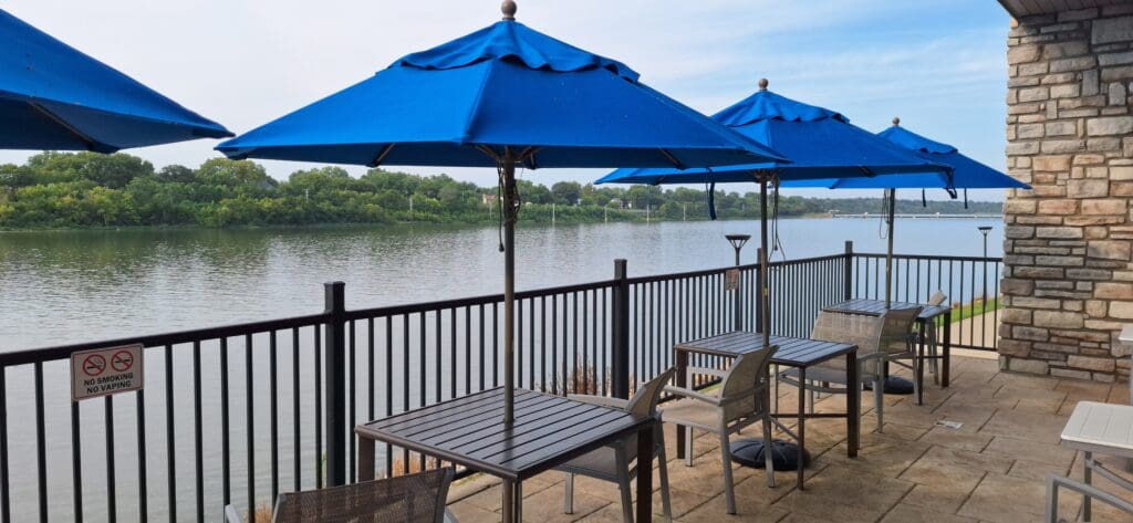 The image shows an outdoor patio area overlooking a body of water. There are several tables and chairs with blue umbrellas providing shade. A metal railing separates the patio from the water, and a "No Smoking No Vaping" sign is visible on the railing. Trees and greenery are visible across the water, and the sky is clear.