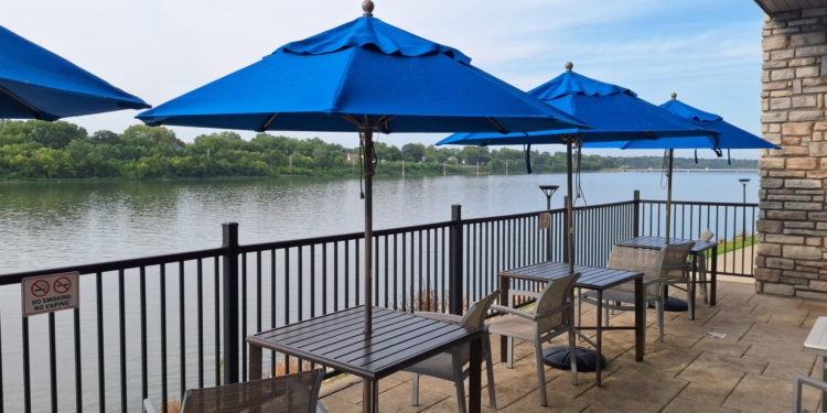 The image shows an outdoor patio area overlooking a body of water. There are several tables and chairs with blue umbrellas providing shade. A metal railing separates the patio from the water, and a "No Smoking No Vaping" sign is visible on the railing. Trees and greenery are visible across the water, and the sky is clear.