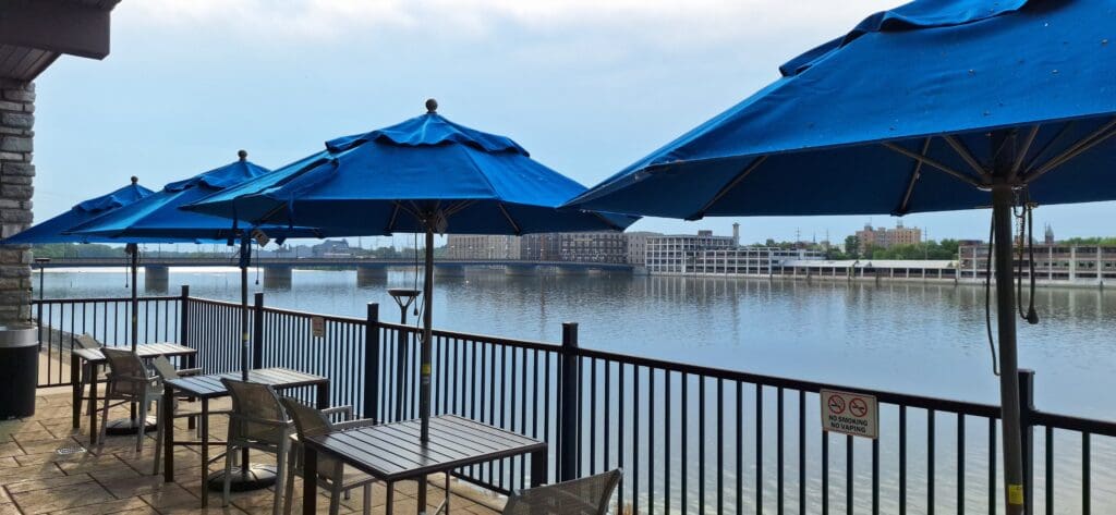 Outdoor seating area by a waterfront, featuring several tables and chairs under large blue umbrellas. A metal railing separates the seating area from the water. In the background, there is a bridge and buildings along the opposite shore. A sign on the railing indicates "No Smoking, No Vaping."