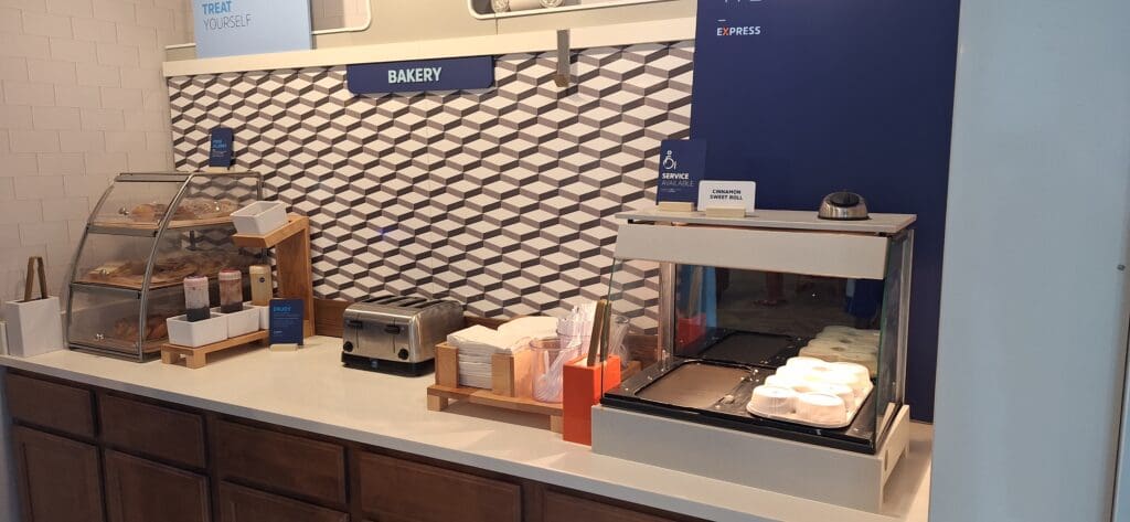 The image shows a bakery counter with a geometric patterned wall in the background. On the counter, there is a display case with various pastries, a toaster, napkins, and utensils. There are also condiment bottles and a sign indicating "Cinnamon Sweet Roll." The area is labeled "Bakery" with a sign above.