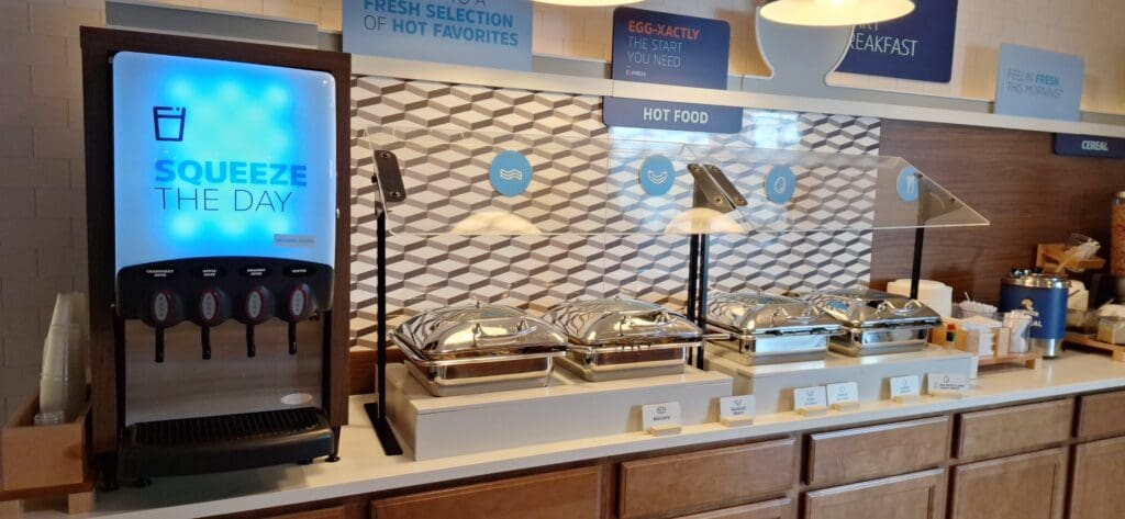 The image shows a breakfast buffet setup. On the left, there is a juice dispenser labeled "Squeeze the Day" with options for cranberry juice, apple juice, orange juice, and water. To the right, there are several covered chafing dishes labeled with food options such as biscuits, sausage gravy, and turkey sausage. Above the buffet, there are signs indicating "Hot Food" and other breakfast-related themes. In the background, there are cereal dispensers and a container labeled "Quaker Oatmeal."