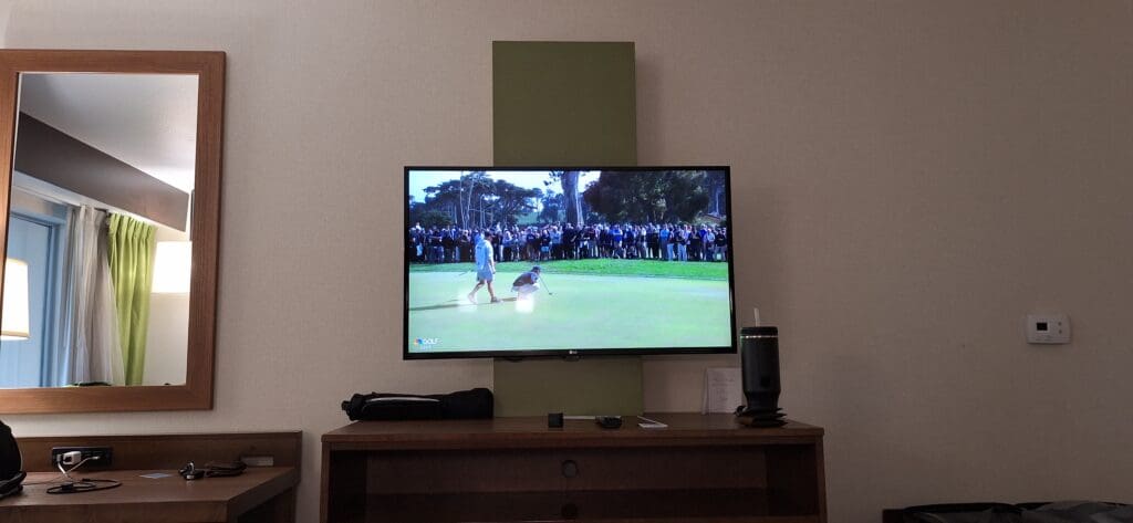 The image shows a hotel room with a television mounted on the wall. The TV is displaying a golf event, with a golfer preparing to putt on a green, surrounded by a crowd of spectators. Below the TV, there is a wooden desk with various items, including a cup with a straw, a remote control, and some electronic devices. To the left, there is a mirror reflecting part of the room, including a lamp and green curtains.