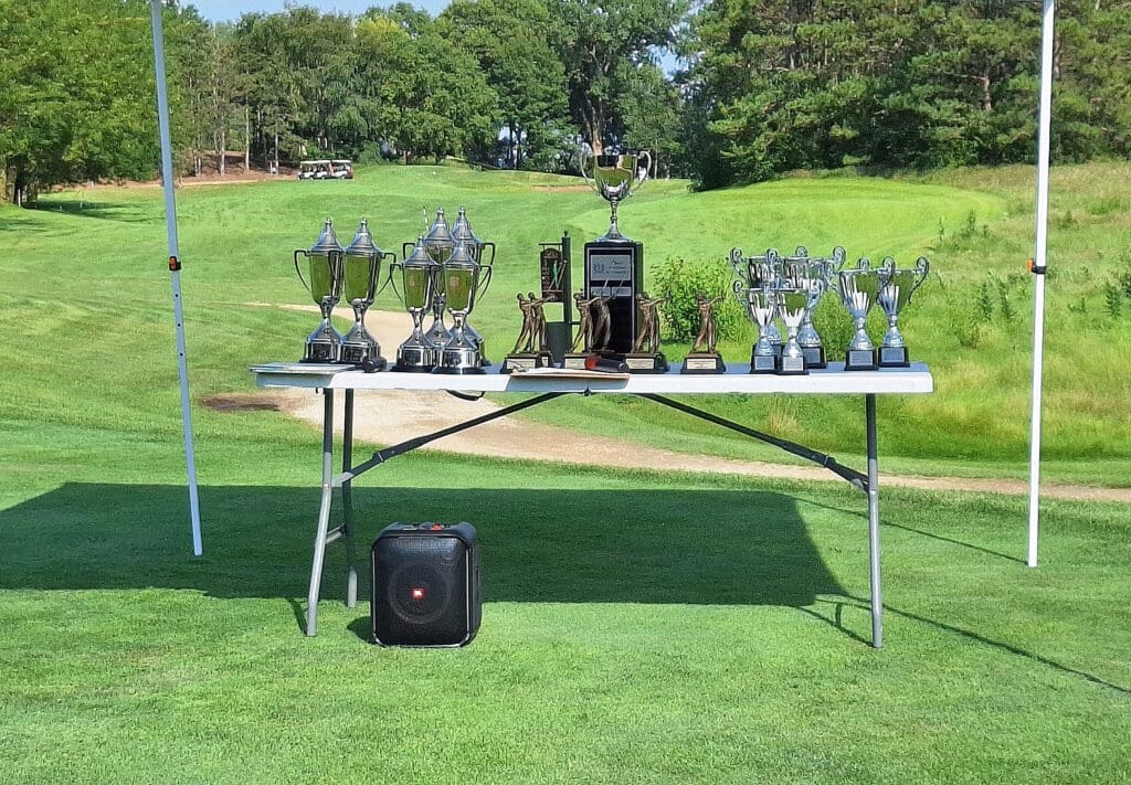 A table is set up outdoors on a grassy area, displaying multiple trophies of various sizes and designs. The background features a golf course with trees and a path. A speaker is placed on the ground in front of the table.