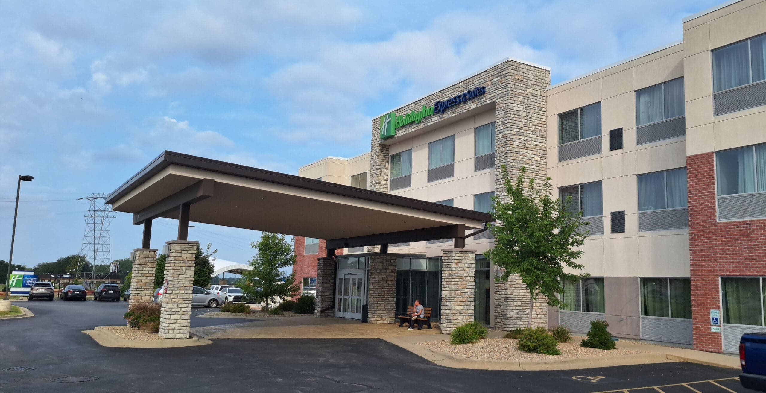 The image shows the exterior of a Holiday Inn Express hotel. The building has a modern design with a combination of stone and brick facade. There is a covered entrance with stone pillars, and a person is sitting on a bench near the entrance. Several cars are parked in the parking lot, and there are trees and landscaping around the area. The sky is partly cloudy.