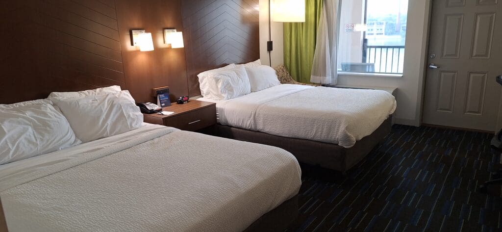 A hotel room with two neatly made double beds featuring white linens. A wooden headboard with two wall-mounted lamps is above the beds. A nightstand between the beds holds a telephone, a clock, and a small sign. A window with green curtains is on the right, and a closed door is visible in the background. The room has a modern and clean appearance.