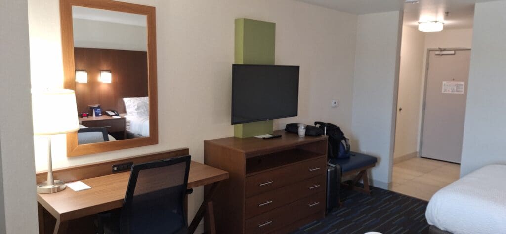 A hotel room with a bed partially visible on the right. A wooden desk with a chair and a lamp is on the left, with a mirror above it reflecting part of the bed and bedside lamps. A flat-screen TV is mounted on the wall above a wooden dresser. There is a small bench with a backpack and a suitcase next to it. The entrance door is visible in the background.
