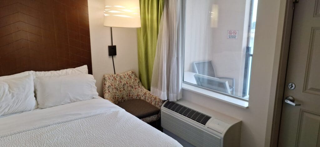 A cozy hotel room corner featuring a neatly made bed with white linens and pillows on the left. Next to the bed is a patterned armchair under a modern lamp with a white shade. A green curtain partially covers a window, through which an outdoor patio with a table and chair is visible. Below the window is an air conditioning unit, and a "No Smoking, No Vaping" sign is on the window.