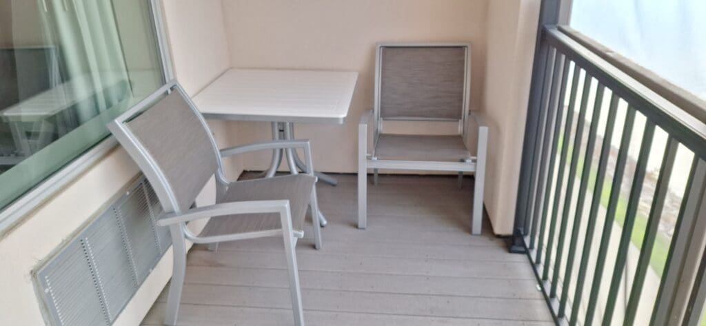 A small balcony with two gray chairs and a white square table. The balcony has a metal railing on one side and a large window on the other. The floor is made of light-colored wood planks.