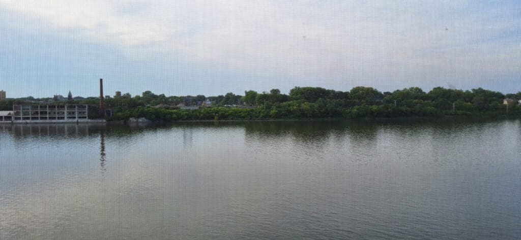A calm river reflecting the sky and surrounding greenery. In the background, there are trees and a few buildings, including a structure with a tall chimney. The sky is partly cloudy, and the scene is viewed through a mesh screen.