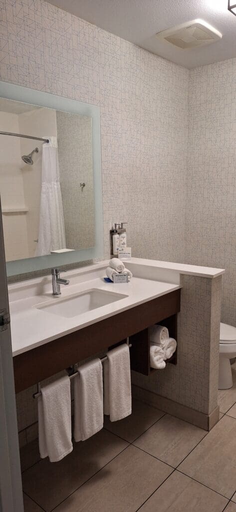 The image shows a modern bathroom with a large mirror above a white countertop sink. There are toiletries and neatly folded towels on the counter. Below the sink, more towels are hanging on a rack. The wall has a geometric pattern, and a shower with a curtain is visible in the background. The floor is tiled, and a toilet is partially visible to the right.