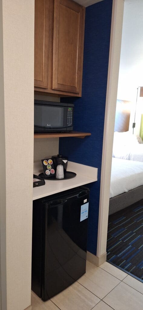 The image shows a small kitchenette area in a hotel room. It includes a black mini-fridge at the bottom, a countertop with a coffee maker and coffee pods, and a microwave on a wooden shelf above. There are wooden cabinets above the microwave. Part of a bed with white bedding is visible in the adjacent room. The walls are a combination of beige and blue.