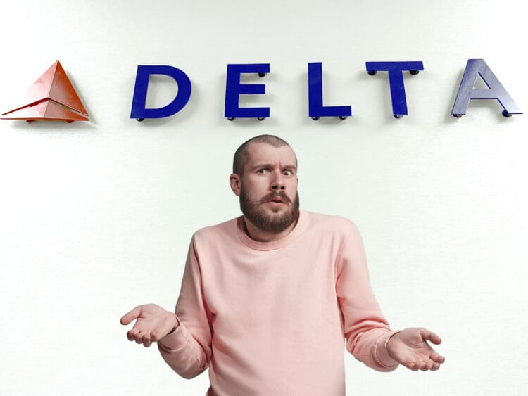 A person with a beard and a puzzled expression is shrugging their shoulders, wearing a pink sweater. They are standing in front of a wall with the word "DELTA" and a red triangular logo.