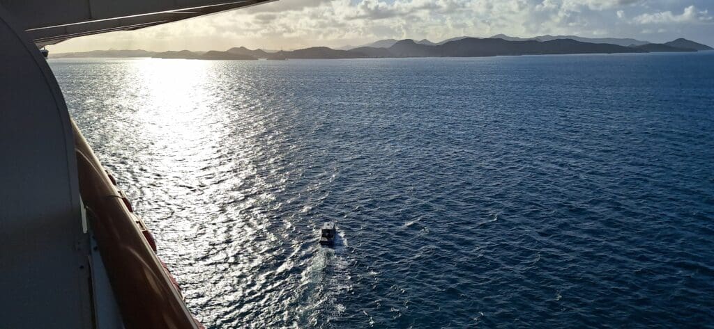 A small boat is sailing on a vast body of water, with the sun reflecting off the surface. In the background, there are distant mountains under a partly cloudy sky. The image is taken from a higher vantage point, possibly from a larger vessel.