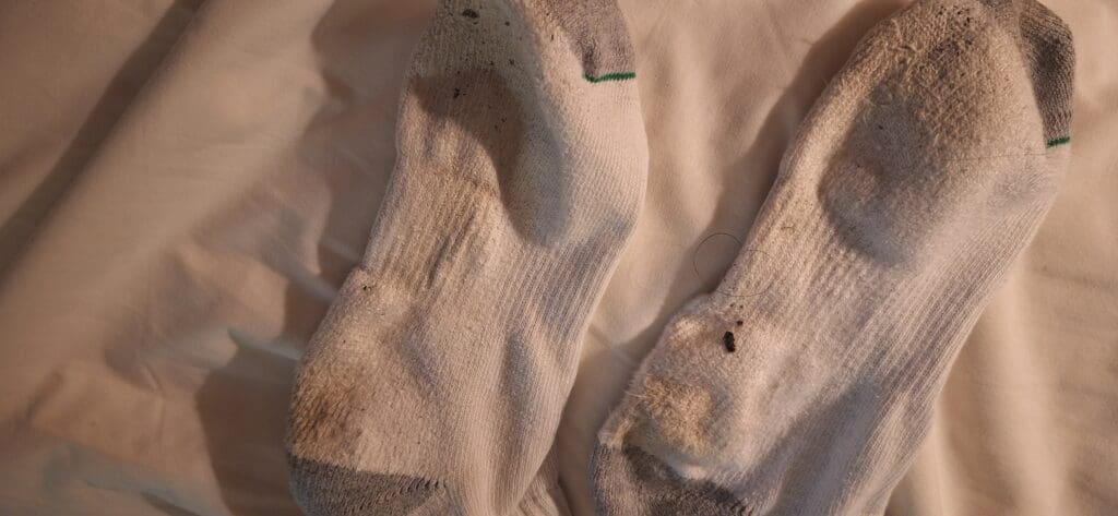 A pair of worn white socks with gray toes and heels, lying on a light-colored fabric surface. The socks have visible dirt marks and some wear and tear.