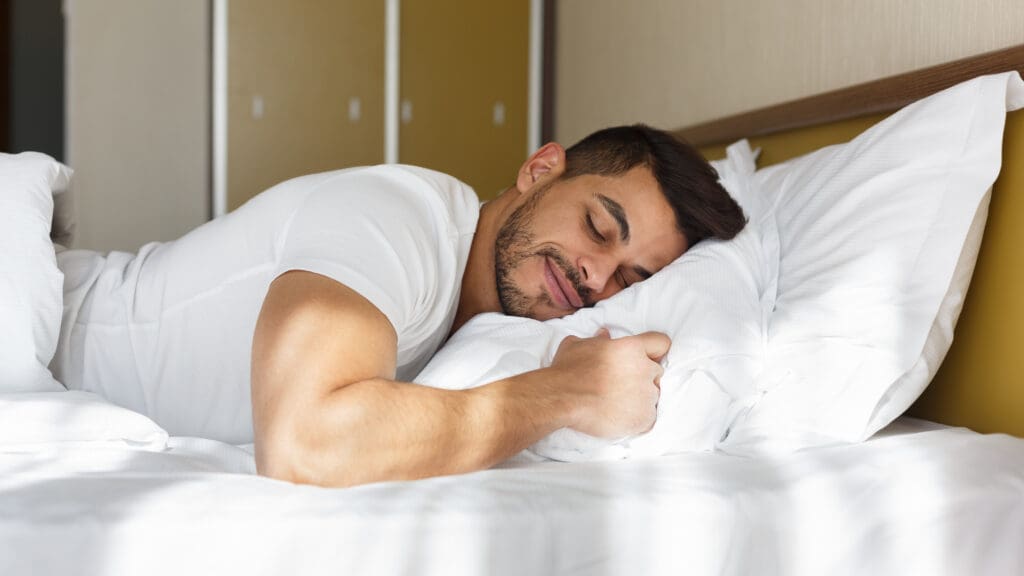 A man is lying in bed, smiling with his eyes closed, and hugging a white pillow. He is wearing a white t-shirt and appears to be relaxed and comfortable. The room has a neutral color scheme with a wooden headboard.