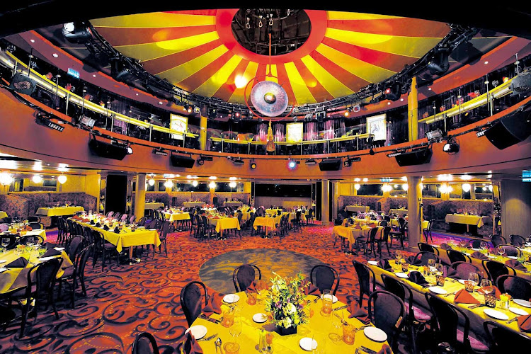 The image shows an elegant, circular dining room with a vibrant, colorful ceiling featuring a large central light fixture. The room is set up with multiple round tables covered in yellow tablecloths, each adorned with floral centerpieces and surrounded by chairs. The space has a two-tiered design with a balcony level above, and the floor is carpeted with a red and orange pattern. The lighting is warm, creating a cozy and inviting atmosphere.