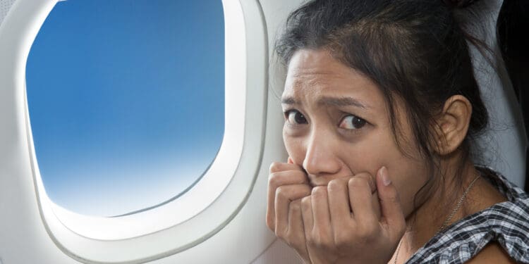 A woman sitting in an airplane seat looks anxious or scared, with her hands near her face. She is next to a window showing a clear blue sky.