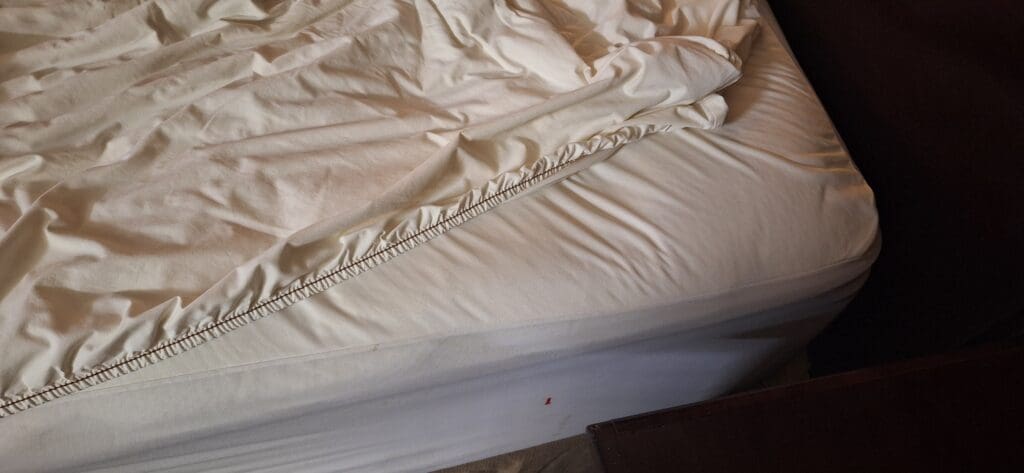 A close-up image of a bed with a white fitted sheet partially pulled back, revealing the mattress underneath. The sheet is slightly wrinkled, and the corner of the bed frame is visible.