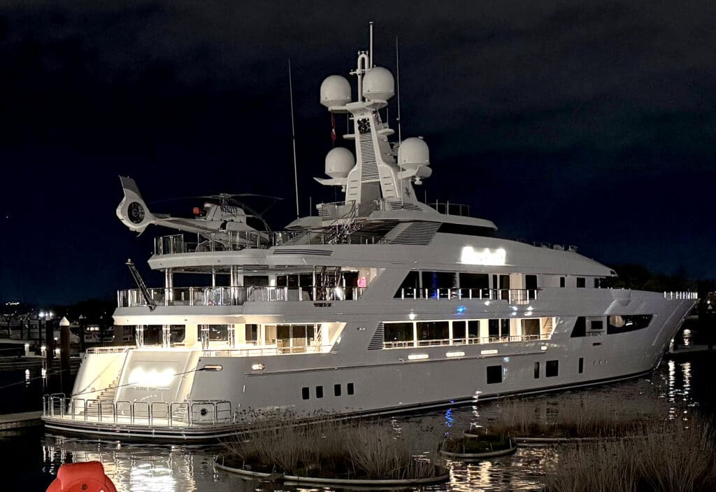 A large, luxurious yacht belonging to Tilman Fertitta is docked at night. The yacht is well-lit, showcasing multiple decks and a sleek design. A helicopter is parked on the top deck. The surrounding water reflects the lights from the yacht, and the sky is dark.