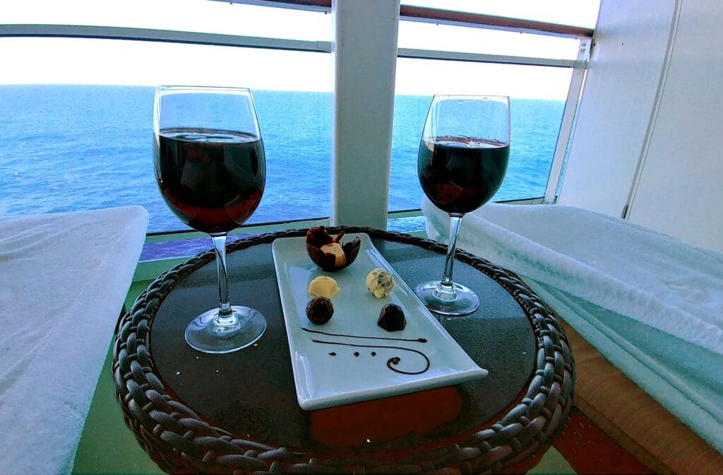 Two glasses of red wine and a plate with assorted chocolates and truffles are on a wicker table. The table is set on a balcony overlooking a vast, calm ocean under a clear sky.