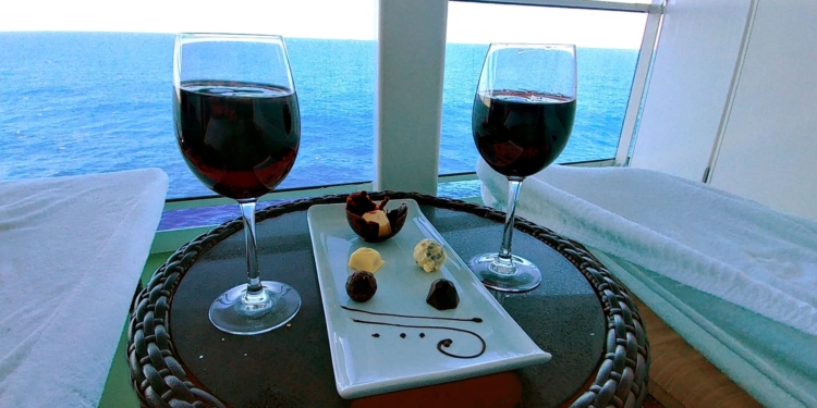 Two glasses of red wine and a plate with assorted chocolates and truffles are on a wicker table. The table is set on a balcony overlooking a vast, calm ocean under a clear sky.