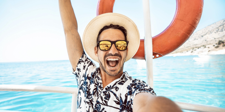 A person wearing a straw hat and sunglasses is taking a selfie on a boat. They are smiling widely and have one arm raised. The person is wearing a patterned shirt. In the background, there is a bright blue sea and a life preserver attached to the boat. The sky is clear and sunny.