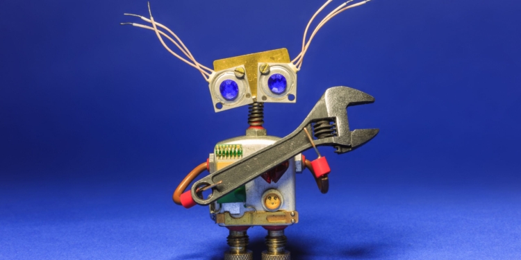 A small robot made from various metal and electronic components is holding a large wrench. The robot has a rectangular body with a circuit board, a head with two large blue eyes, and thin wires protruding from the top. It stands on a blue surface with a blue background.