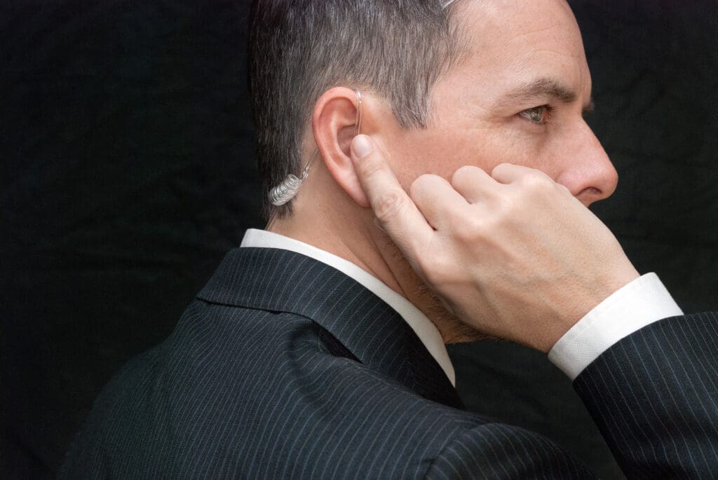 A man in a suit is shown from the side, wearing an earpiece with a coiled wire, suggesting he might be a security agent or bodyguard. He is touching his ear, possibly listening to a communication device. The background is dark.
