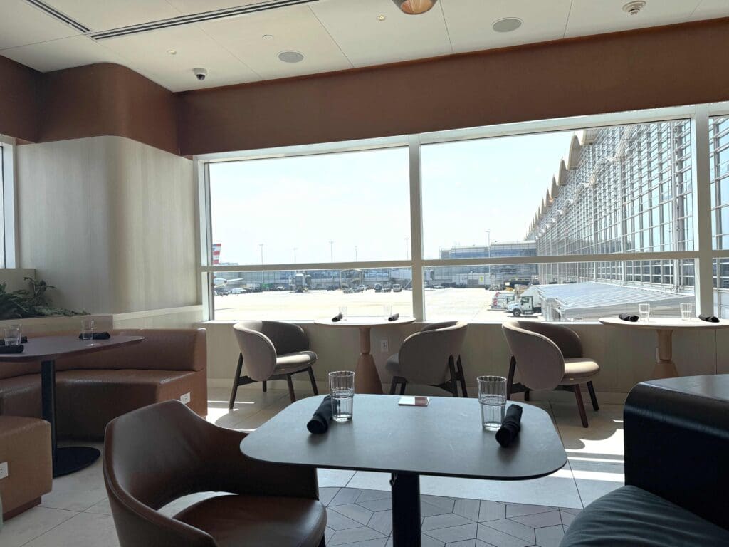 The image shows a modern airport lounge with several tables and chairs arranged near large windows. The windows offer a view of the airport tarmac and a terminal building with a distinctive wavy roof. Each table has a glass of water and a rolled black napkin. The interior features a mix of brown and beige tones, with some greenery visible in the corner. The space is well-lit with natural light coming through the windows.
