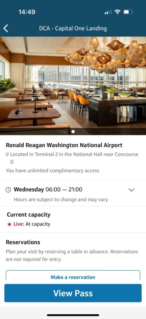 The image shows a mobile app screen for "DCA - Capital One Landing" at Ronald Reagan Washington National Airport. It features a photo of a modern lounge area with tables, chairs, and decorative lighting. The text indicates the lounge is located in Terminal 2, National Hall near Concourse D, with unlimited complimentary access. The hours are 06:00 to 21:00 on Wednesday. The current capacity is "At capacity." There is an option to make a reservation, though it is not required for entry. A "View Pass" button is visible at the bottom.