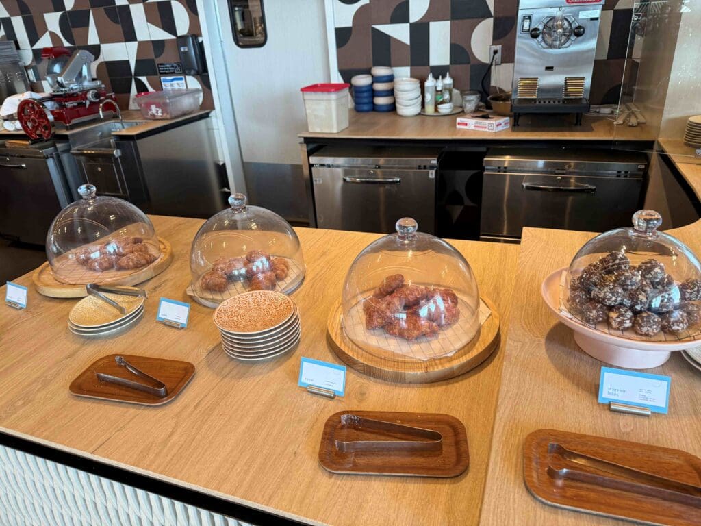 The image shows a countertop display in a bakery or café setting. There are three glass domes covering different types of pastries. The first dome contains croissants, the second has sugar-coated pastries, and the third holds chocolate-covered treats. In front of each dome, there are small signs and tongs on wooden trays. Behind the counter, there are kitchen appliances, plates, and utensils. The background features a geometric patterned wall.