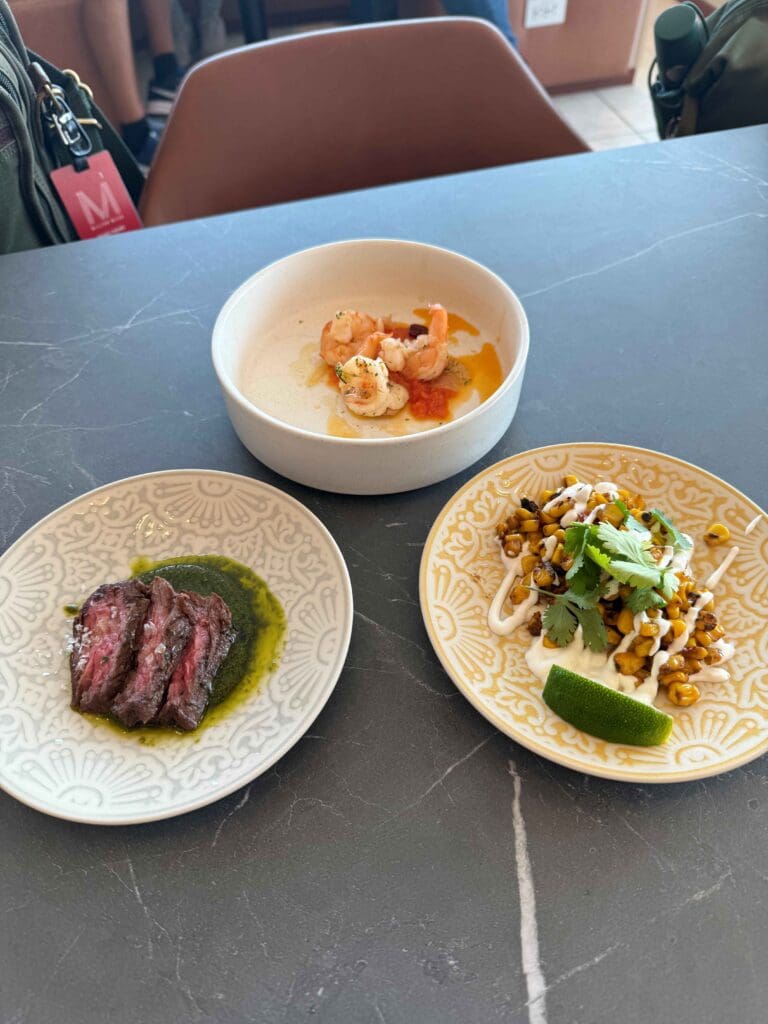 Three dishes are arranged on a dark table. The top dish contains shrimp in a light sauce. The bottom left dish has slices of steak with a green sauce. The bottom right dish features roasted corn topped with cilantro and a drizzle of white sauce, accompanied by a lime wedge.