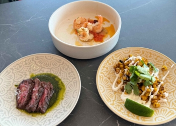 Three dishes are arranged on a dark table. The top dish contains shrimp in a light sauce. The bottom left dish has slices of steak with a green sauce. The bottom right dish features roasted corn topped with cilantro and a drizzle of white sauce, accompanied by a lime wedge.