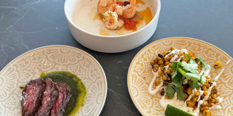 Three dishes are arranged on a dark table. The top dish contains shrimp in a light sauce. The bottom left dish has slices of steak with a green sauce. The bottom right dish features roasted corn topped with cilantro and a drizzle of white sauce, accompanied by a lime wedge.