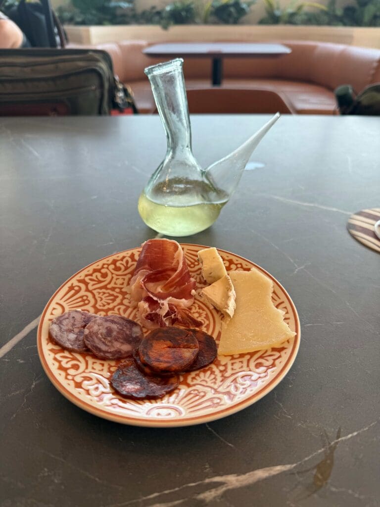 The image shows a plate with a variety of cured meats and cheeses, including slices of salami, chorizo, prosciutto, and two types of cheese. The plate has an ornate design. In the background, there is a glass vessel with a spout containing a light-colored liquid, possibly wine. The setting appears to be a dining area with a table and seating.