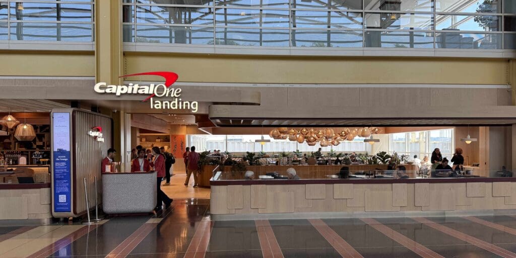 The image shows the entrance to a Capital One Landing lounge in an airport. The lounge has a modern design with a sign displaying the Capital One logo. Inside, there are people sitting and interacting in a well-lit area with hanging lights and plants. The space has large windows allowing natural light to enter.