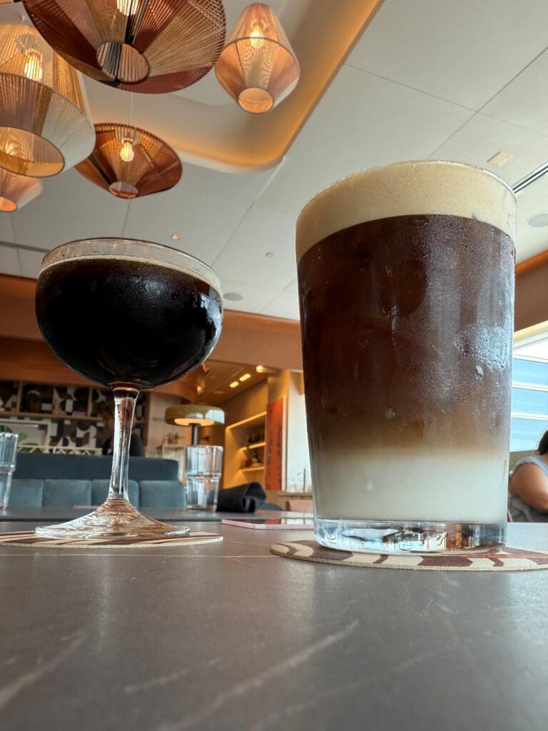 The image shows two drinks on a table in a restaurant setting. On the left is a dark beverage in a coupe glass, and on the right is a layered iced coffee drink in a tall glass, with a creamy layer at the bottom and a frothy top. The background features modern decor with hanging light fixtures and a glimpse of the restaurant interior.