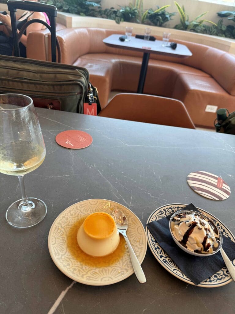 The image shows a table with a dessert setup. There is a glass of white wine on the left, a plate with a flan and a spoon in the center, and a bowl of ice cream drizzled with chocolate sauce on the right, placed on a decorative plate with a napkin. In the background, there is a seating area with brown leather couches, a table with glasses, and some greenery. A suitcase and a bag are visible near the seating area.