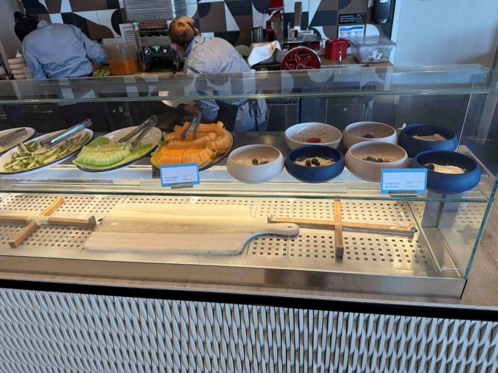 The image shows a food display counter with various items. On the left, there are plates with sliced fruits, including green melon and cantaloupe. On the right, there are several bowls containing yogurt and granola, topped with berries. Behind the counter, two people are preparing food. The counter has a wooden cutting board and a decorative pattern on the front.