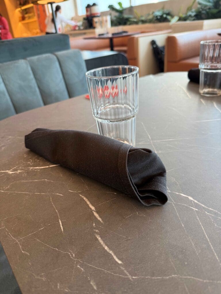 A clear glass and a rolled-up black napkin are placed on a dark marble table. In the background, there are cushioned seats and a person standing near a counter with plants visible nearby.