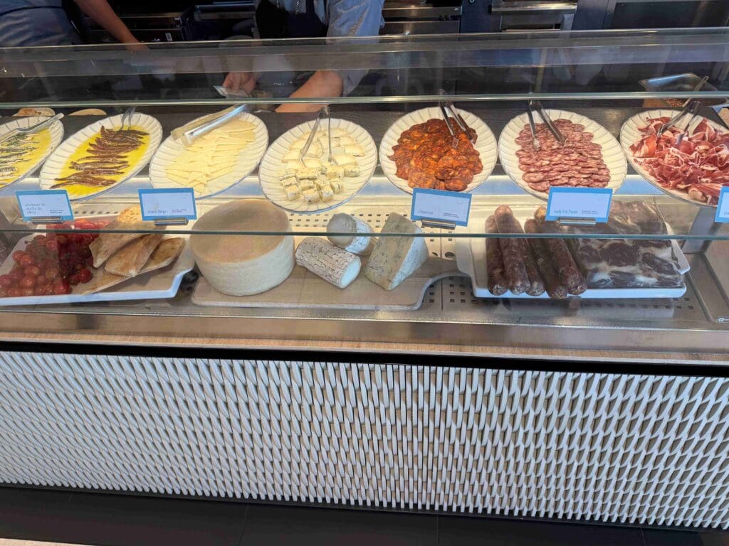 The image shows a deli counter display with various types of sliced meats and cheeses. There are plates with anchovies, sliced cheese, cubes of cheese, chorizo, salami, and prosciutto. Below the plates, there are whole cheeses, bread, cherry tomatoes, and grapes. The items are labeled with small signs, and there are tongs for serving.