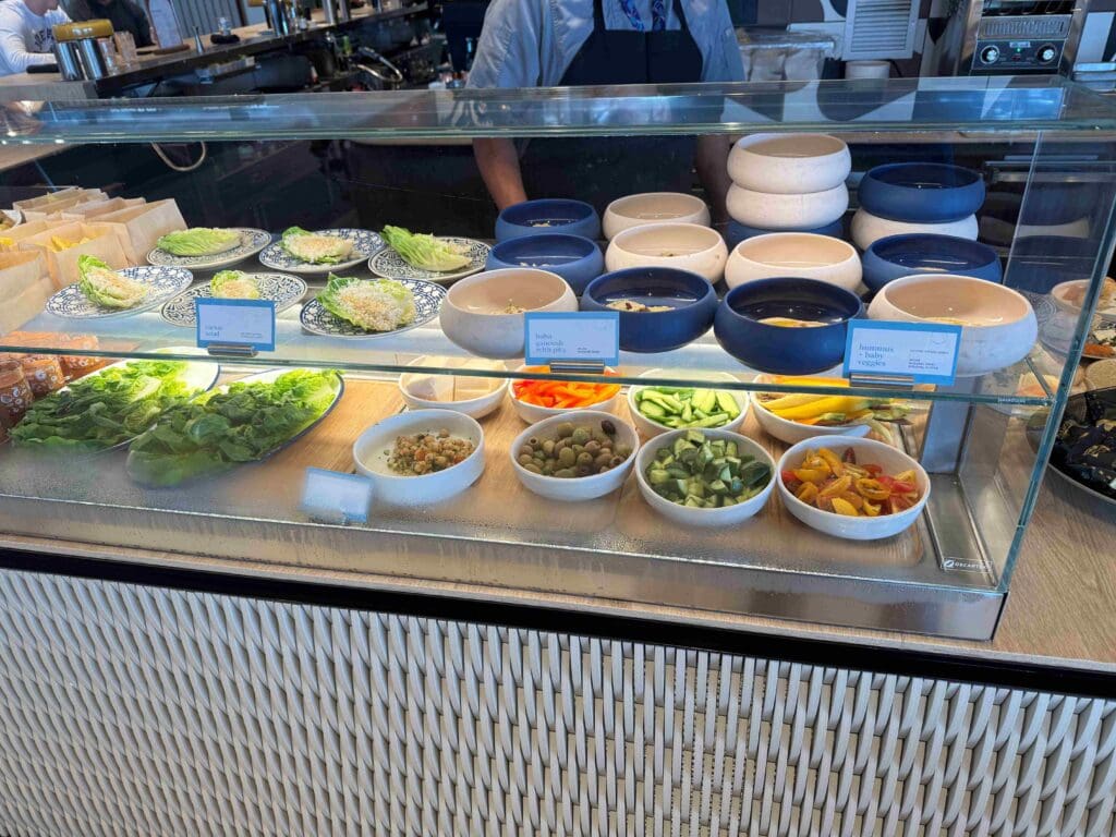 The image shows a display case in a restaurant or café setting. Inside the case, there are various food items arranged on two shelves. The top shelf has plates with lettuce wraps and several stacked bowls, some of which are blue and others are white. The bottom shelf contains bowls with different ingredients, including sliced cucumbers, olives, chickpeas, and colorful bell peppers. There are also plates with leafy greens. Small signs with labels are placed in front of some items.
