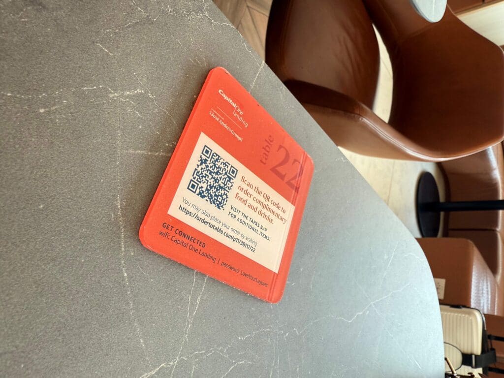 The image shows a table with a red card labeled "table 22" on a gray surface. The card includes a QR code and text instructing users to scan it to order complimentary food and drinks. It also provides a URL for additional items and Wi-Fi connection details. In the background, there are brown chairs and part of a suitcase is visible.