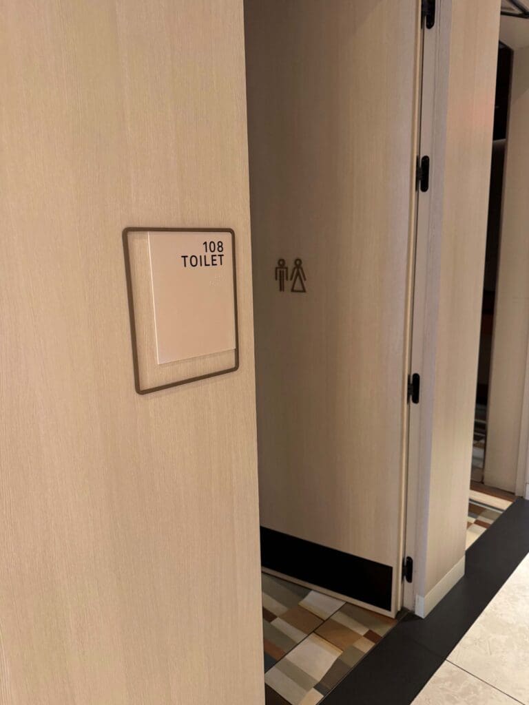 The image shows a light-colored wooden door with a sign that reads "108 TOILET" in black text. Below the text, there is Braille. The door is slightly open, revealing a tiled floor inside. On the door, there are symbols indicating a unisex toilet.