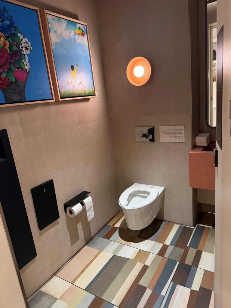 The image shows a modern bathroom with a wall-mounted toilet. The floor has multicolored striped tiles. On the wall, there are two framed artworks: one features a person with a floral headdress, and the other depicts a person flying a kite in a field. A round light fixture is mounted on the wall above the toilet. A toilet paper holder with a roll is attached to the wall.