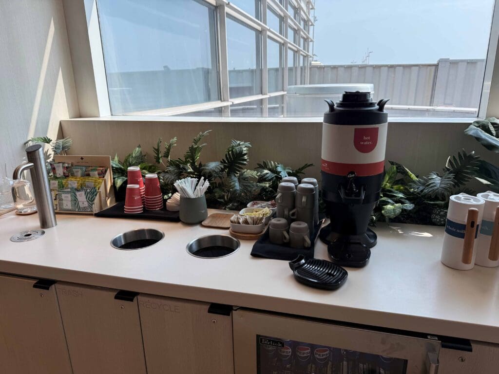 The image shows a beverage station with a large hot water dispenser labeled "hot water" on a counter. There are stacks of red paper cups, a container of stir sticks, and various tea bags displayed in a box. Several gray mugs are placed next to the dispenser. The counter also has built-in trash and recycle bins. In the background, there are decorative green plants and a large window with a view of the outside.