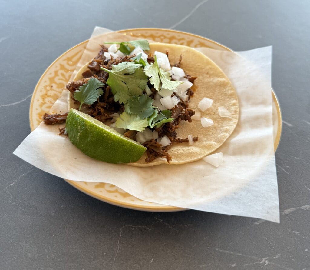 A taco on a decorative plate with a corn tortilla, topped with shredded meat, chopped onions, and cilantro. A lime wedge is placed on the side. The plate is lined with a piece of parchment paper.