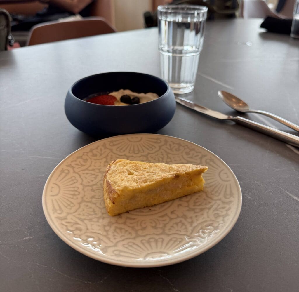 A slice of omelette is on a decorative white plate in the foreground. In the background, there is a bowl with yogurt topped with strawberries and blueberries. A glass of water, a fork, and a spoon are placed on the table next to the bowl.