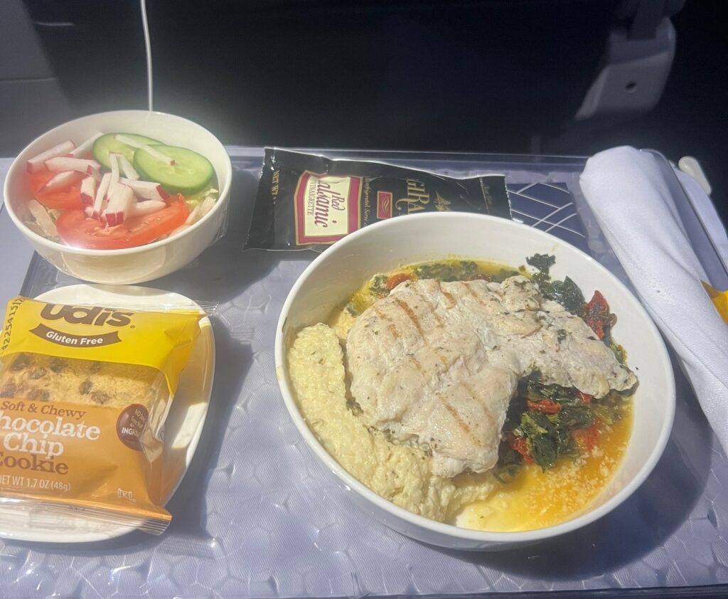 The image shows an airplane meal on a tray table. There is a bowl with grilled chicken, mashed potatoes, and vegetables. Next to it is a small salad with cucumber, tomato, and radish slices. A packet of balsamic vinaigrette dressing is also visible. Additionally, there is a packaged gluten-free chocolate chip cookie and a rolled-up napkin with cutlery.